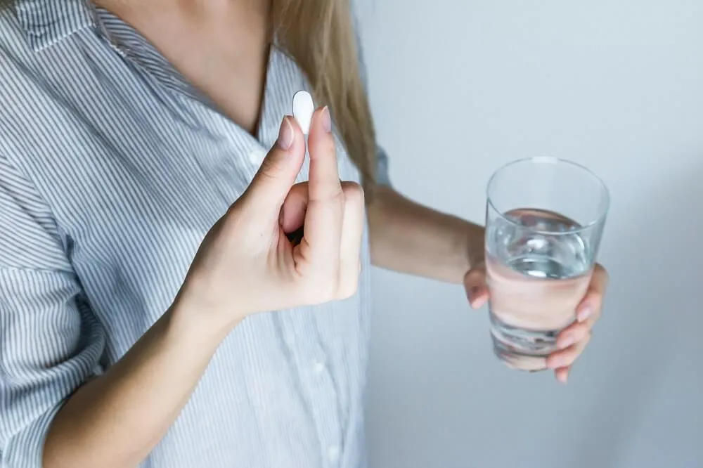 穿淺藍條紋襯衫的女性一手捏著一顆白色藥錠、另一手拿著裝水的玻璃杯，畫面只拍到胸口與雙手