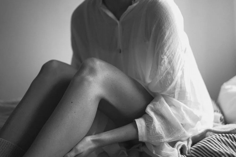 黑白照，穿寬鬆白襯衫的女性坐在床上，雙腿彎曲交疊，一手輕放在小腿上，畫面聚焦在膝蓋